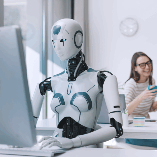 Ein humanoider Roboter mit weiblichen Merkmalen arbeitet konzentriert an einem Computer in einem modernen Büro, im Hintergrund sitzt eine lachende Frau mit Smartphone.