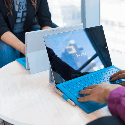 Zwei Personen arbeiten an Microsoft Surface Tablets mit blauer Tastatur an einem runden Tisch in einem modernen Büro.