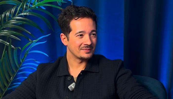 Männlicher Host in schwarzem Outfit während eines Interviews auf einem blauen Sessel mit Hintergrund aus Pflanzen und Vorhang